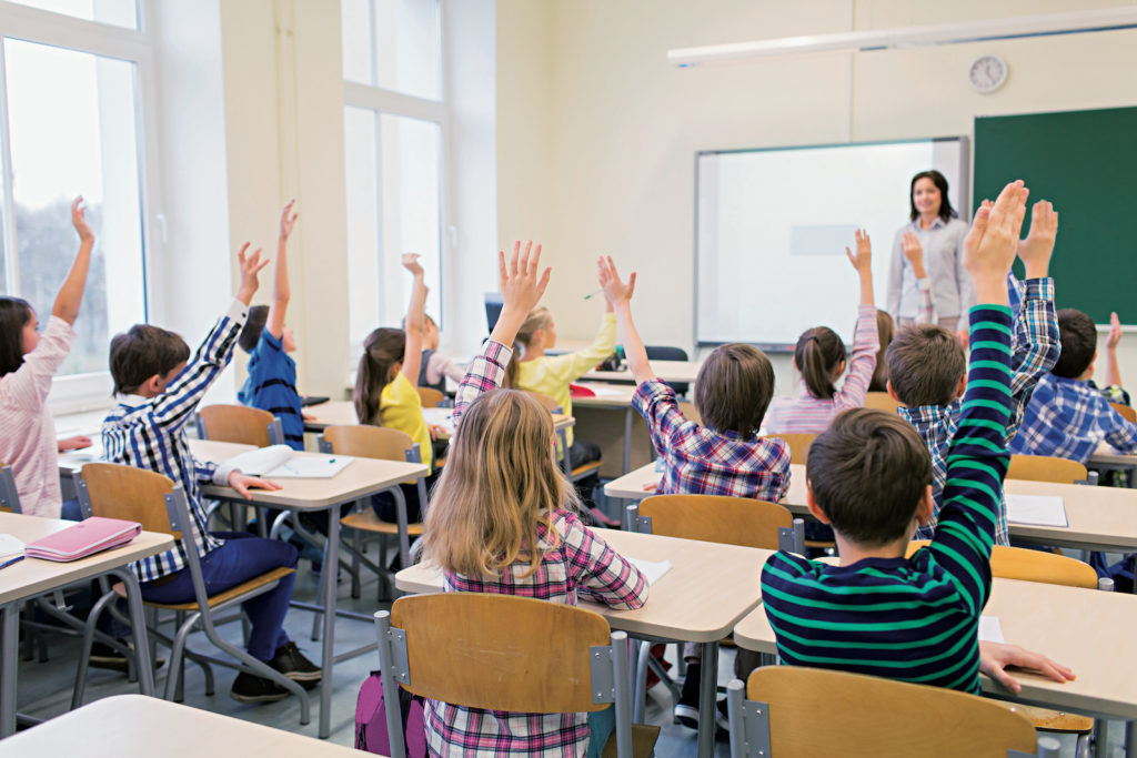 group of school kids raising hands in classroom